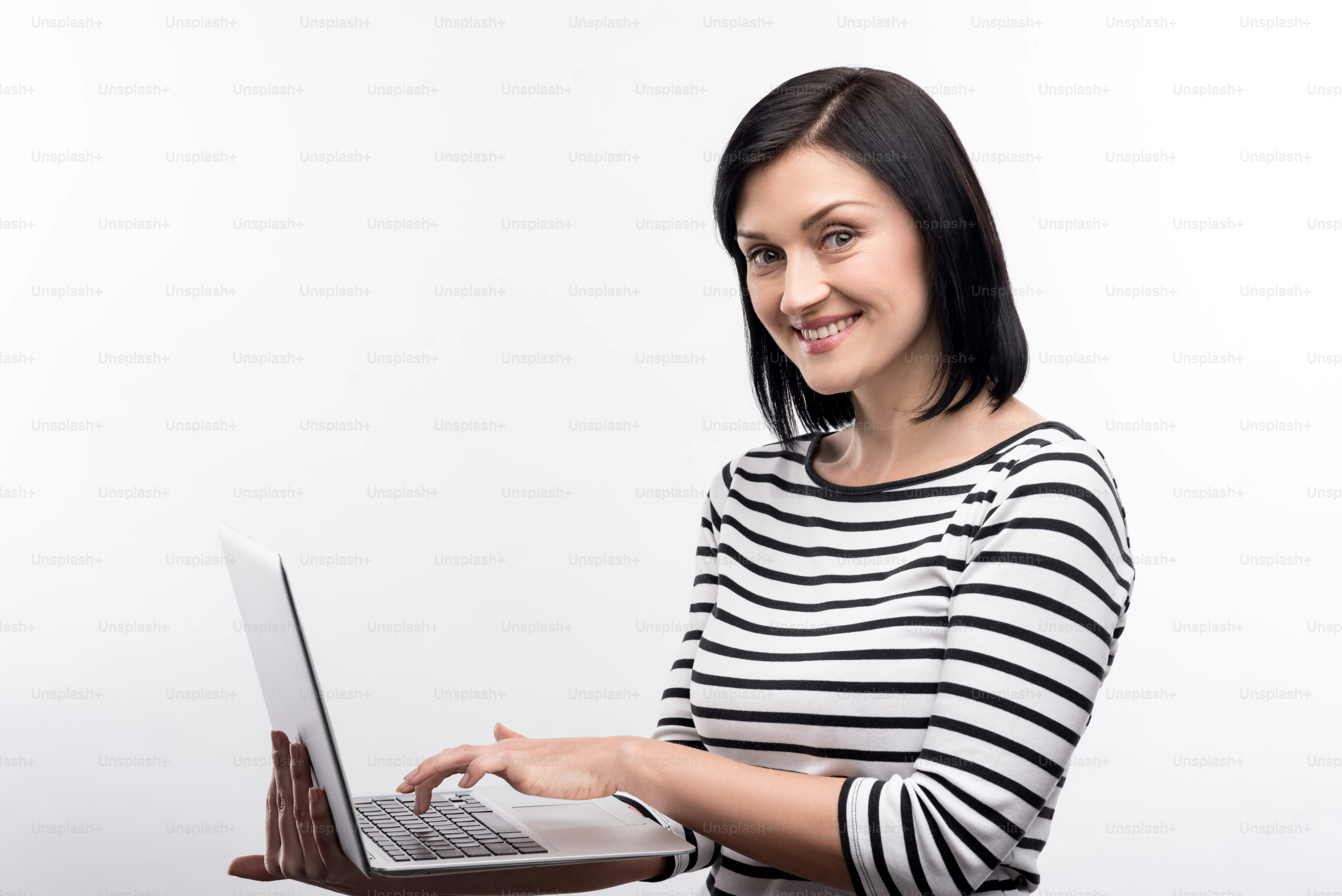 Love my work. Pretty dark-haired woman in a striped pullover standing half-turned and posing for the camera while working on a laptop